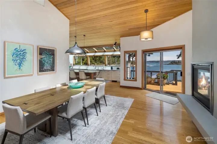 Open-concept dining area flows from the kitchen to the living space, with vaulted ceilings, warm wood details, a modern fireplace, and direct access to waterfront deck living.