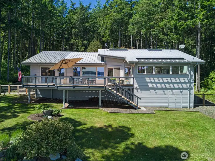 Rear exterior view highlights the spacious deck, glass railing, updated outdoor living areas, and private lawn overlooking the waterfront and surrounding forest.