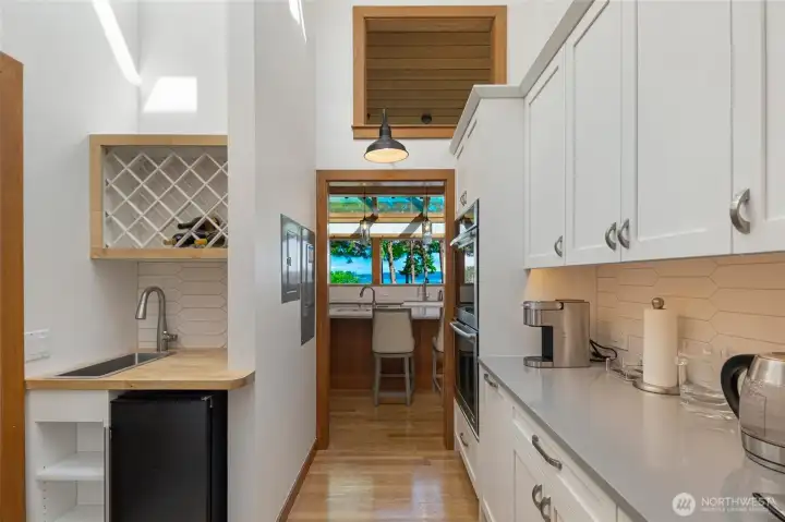 Functional galley-style secondary kitchen or wet bar area with cabinetry, sink, wine storage, and a view connection toward the main kitchen and waterfront beyond.