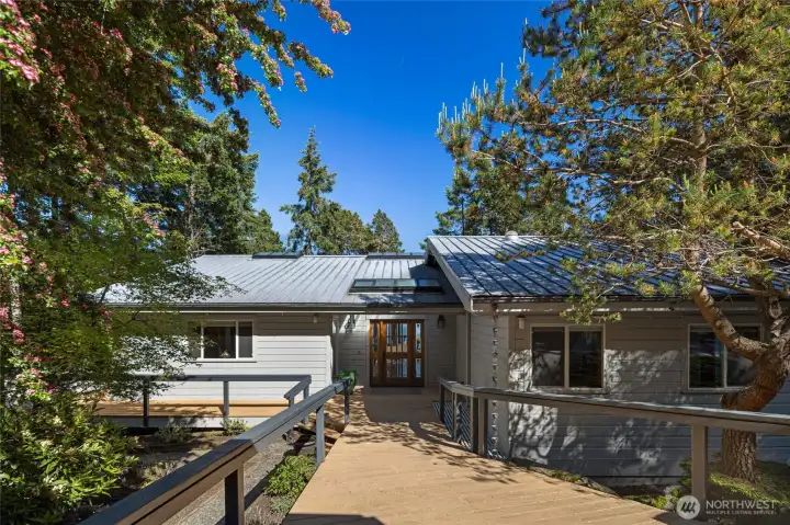 Welcoming entry deck with natural wood details, mature landscaping, and forested surroundings creates a warm arrival to this private waterfront home.