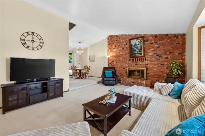 Spacious living room with vaulted ceilings, striking brick fireplace, and open flow to the dining area. A warm and welcoming space ideal for relaxing or entertaining guests.