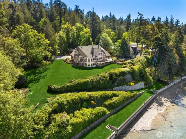 This breathtaking shot showcases a  magnificent waterfront estate on Whidbey  Island, spanning over 7 lush acres. The  expansive 6,120 sq ft home, nestled amidst  verdant landscapes, boasts six luxurious  bedrooms and offers 254 feet of private  waterfront.