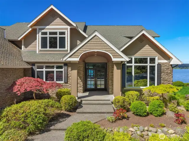 The front facade of the Whidbey Island estate  showcases exceptional architectural design  harmoniously blending with its natural  surroundings. The grand entrance is  highlighted by dramatic gabled rooflines and  expansive windows that invite abundant  natural light.