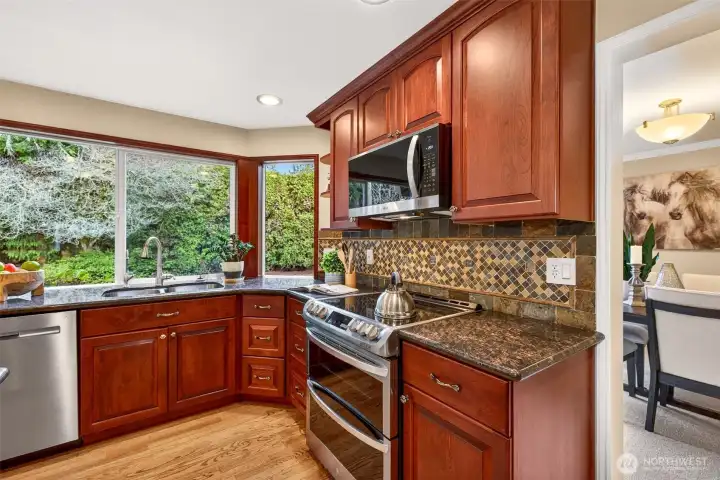 Inviting, updated kitchen featuring rich cabinetry, granite counters, mosaic tile backsplash, stainless appliances, and a large window at the sink with a view to the fenced backyard.