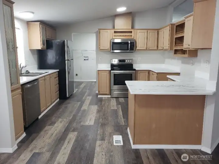 Picture of the Huge Kitchen Showing Ample Cabinet Space, Newer Counter Tops, LED Lighting, All Stainless Appliances Including Built In Microwave, Glass Top Stove Oven, Trim, and Vinyl Plank Flooring.
