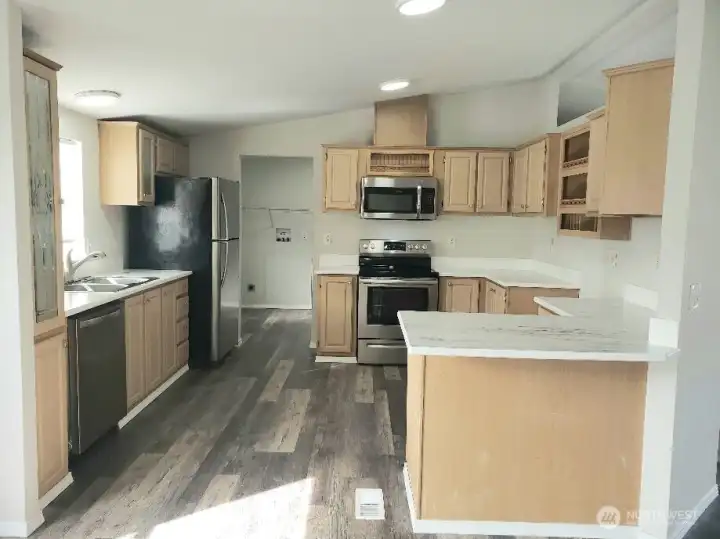 Picture of the Huge Kitchen Showing Ample Cabinet Space, Newer Counter Tops, LED Lighting, All Stainless Appliances Including Built In Microwave, Glass Top Stove Oven, Trim, and Vinyl Plank Flooring.
