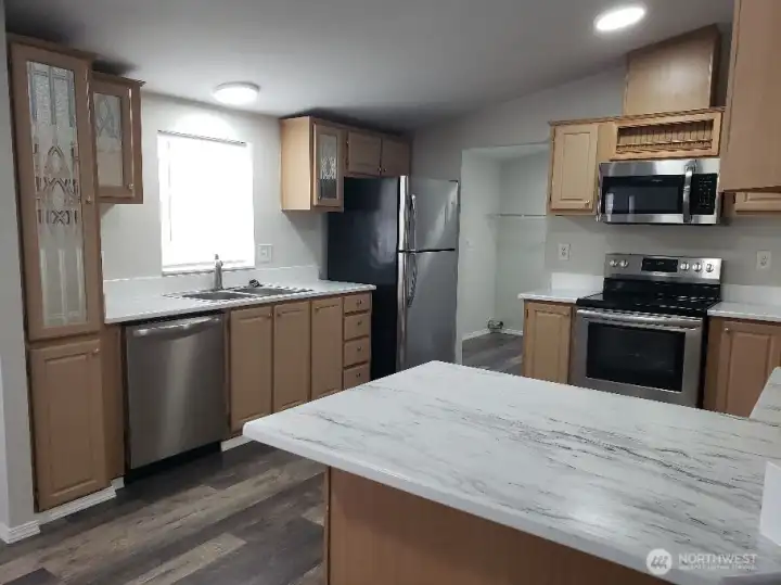 Another Picture of the Huge Kitchen Showing Ample Cabinet Space, Newer Counter Tops, LED Lighting, All Stainless Appliances Including Built In Microwave, Glass Top Stove Oven, Trim, and Vinyl Plank Flooring.