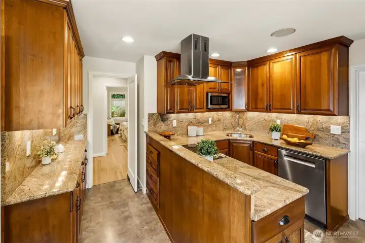 Another view of the kitchen with granite slab counter tops and more storage. Through the doorway find 3 bedrooms, main level baths & hallway to the lower level.