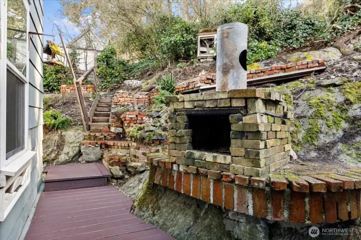 A rustic outdoor fireplace is nestled into the hillside, with brick terraces stepping upward from it along the slope.