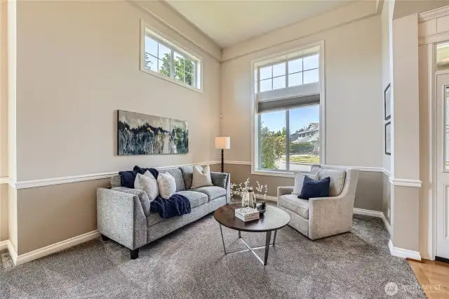 This inviting living room exudes elegance and comfort. Bathed in natural light from tall windows and a charming transom, the space features soaring ceilings that enhance its airy and open feel. The neutral-toned walls and plush carpeting create a warm and sophisticated backdrop, while stylish furnishings—including a tufted sofa, modern armchair, and sleek coffee table—add a contemporary touch. Perfect for entertaining or relaxing, this beautifully staged room offers a welcoming ambiance with picturesque views of the front yard.