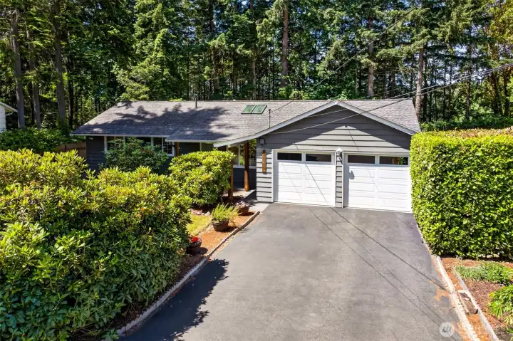 The attached two-car garage and extended driveway provide generous off-street parking and storage, framed by mature hedges that screen the approach for added seclusion.