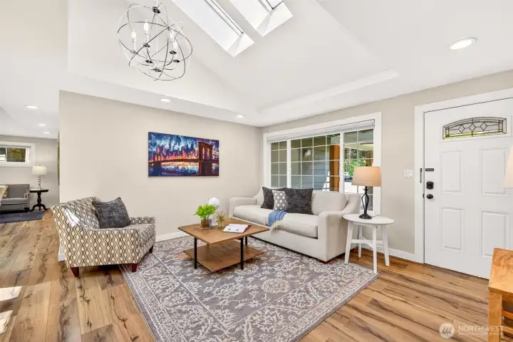 The bright living area centers on a sculptural chandelier and a wide window that frames the front garden, offering a comfortable, light-filled space to gather and unwind.