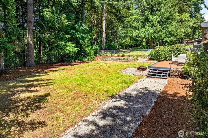 The expansive backyard offers a level lawn, gravel pathways, and terraced garden beds, with the composite deck and fire pit creating distinct zones for gardening, gathering, and quiet relaxation.