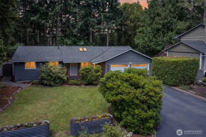 As evening settles, warm light spills from the windows of this single-level home, highlighting its inviting curb appeal, mature hedging, and private setting against the trees.