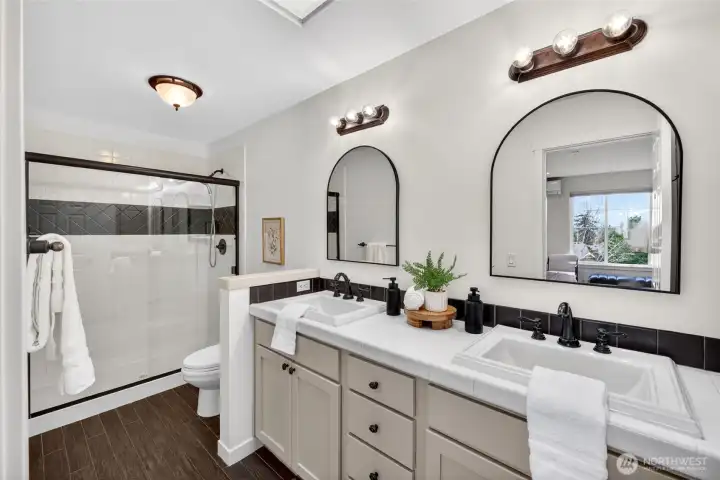Spacious primary bath designed for both comfort and functionality, featuring dual vanities with ample counter space and storage. A large walk-in shower with tile surround and glass enclosure adds a clean, updated feel, while the thoughtful layout provides separation and ease of use for daily routines. Bright, well-balanced lighting and a connection to the adjacent bedroom complete the suite-like experience.