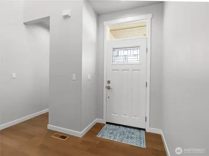 Step inside this welcoming soaring entryway featuring a classic craftsman-style front door with decorative leaded glass insert and transom window above, bringing in natural light. Warm engineered hardwood-style flooring and clean neutral tones set the tone for the rest of the home.