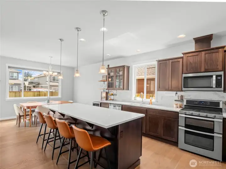 A chef's dream kitchen featuring rich dark-stained cabinetry, quartz countertops, a large center island with seating for four, pendant lighting, and stainless steel appliances including a double oven range. Under-cabinet lighting adds both ambiance and function to this beautifully designed space.