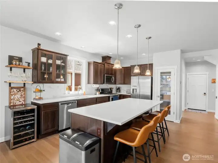 The kitchen's perimeter features generous quartz counter space, a deep double undermount sink, tile backsplash, and matching dark cabinetry with ample storage. The layout is designed for both efficiency and style, with a clear sightline to the dining area and backyard.