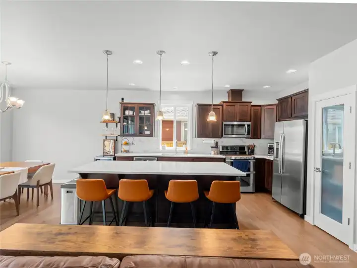 Wide view of the kitchen and adjoining dining space showcases the open, airy floor plan with recessed lighting, pendant fixtures, stainless appliances, and beautiful engineered hardwood flooring that ties the spaces together effortlessly. Also shown is the kitchen pantry.