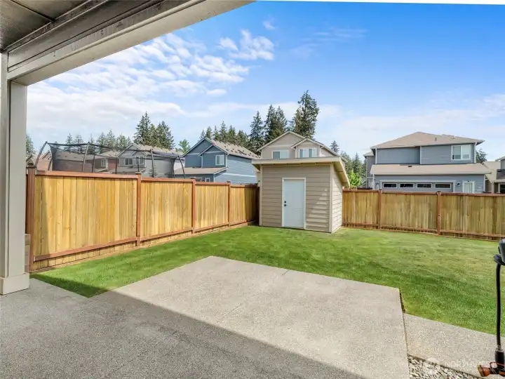 Wide view of the fully fenced backyard showcases the custom-built shed — perfectly matching the home's exterior — set against a well-maintained lawn and covered patio. The shed provides excellent additional storage for tools, gear, or hobbies.