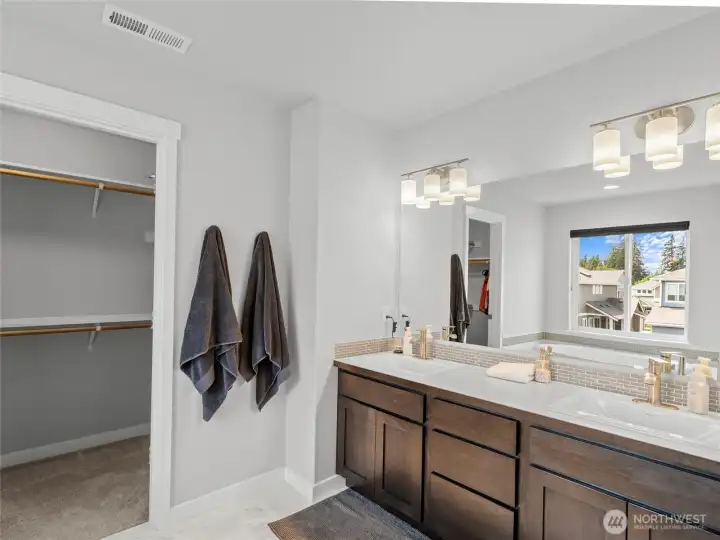The luxurious primary bathroom features dual sinks set in a rich dark-stained vanity with quartz countertops, tile backsplash, gold-tone fixtures, and direct access to the walk-in closet. Natural light and modern lighting fixtures complete this spa-inspired space.