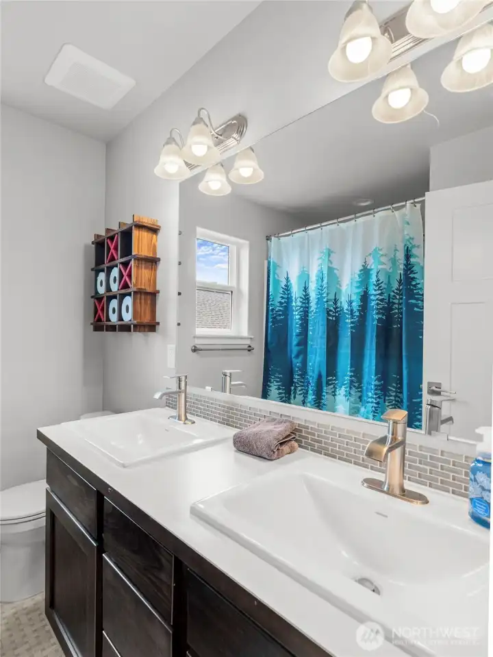 The full upstairs bathroom is well-appointed with a double-sink vanity, dark-stained cabinetry, tile backsplash, and a shower/tub combo. Plenty of counter space makes this bathroom ideal for sharing.