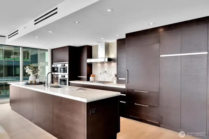 An eating bar is located at the far end of the kitchen island, close to the coffee/espresso machine and the wine/beverage fridge.