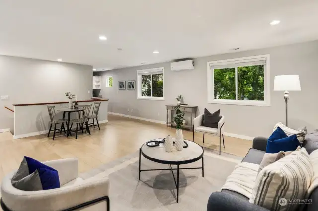 The airy upstairs living room, flooded with natural light, offers serene views of gardens and trees. Recessed lighting complements the light-colored walls, creating a warm ambiance.