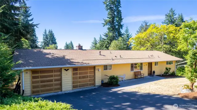 This welcoming exterior view highlights the home’s inviting curb appeal. The yellow siding paired with warm wood garage doors creates a cheerful, classic look. A wide driveway offers plenty of parking, while the covered entry and sitting area add a comfortable, approachable feel. Surrounded by greenery and mature landscaping, the home strikes a balance between convenience and character. It’s a great example of a well-kept property with room to personalize and make your own.
