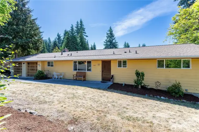 This full exterior shot highlights the home’s cheerful yellow siding, complemented by wood-trimmed details and a matching garage. The paved front walkway and tidy landscaping create curb appeal, while the long layout emphasizes one-level living. Surrounded by tall evergreens, the home feels private yet connected to its natural setting—a perfect blend of suburban living and PNW charm.