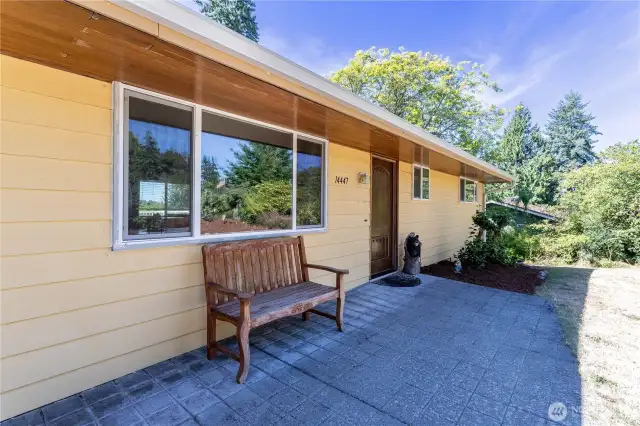 The front entry is simple and inviting, with a paved sitting area that sets a welcoming tone. A wooden bench provides a charming touch, while the mature landscaping adds greenery and shade. It’s a straightforward but cozy entrance that reflects the home’s approachable style.