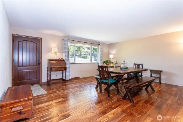This main living space features an open layout with updated hardwood floors and plenty of natural light. A large front window frames the greenery outside while creating a bright, welcoming atmosphere inside. The dining area flows naturally into the living space, making it ideal for gatherings. Neutral walls and warm flooring provide the perfect backdrop for any style, whether modern, rustic, or traditional. This room sets the tone for the home—comfortable, spacious, and full of potential.