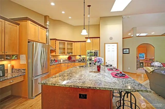 Kitchen view showing work desk, French door refrigerator, granite island with dishwasher, wall ovens and pantry.  Archway leads to home office.