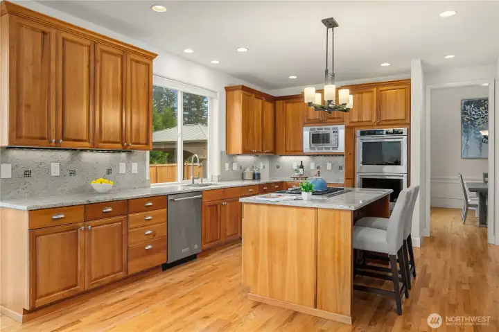 This view of the kitchen shows the flow from the dining room, the eating space on the island, the beautiful granite counters and backsplash highlighted by the under counter lights, and the large kitchen window over the sink.