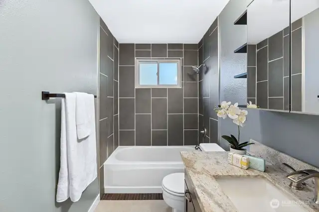 This beautifully updated bathroom features sleek floor-to-ceiling tile in the shower/tub combo, a modern vanity with granite countertops, and elegant fixtures throughout. A large mirror and built-in shelving add both style and storage, while natural light from the window brightens the space, creating a fresh and relaxing atmosphere.