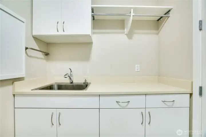 Opposite the laundry zone, a built-in utility sink and extended countertop workspace offer endless possibilities—from crafting and gardening prep to pet care or project organization—all supported by abundant upper and lower cabinetry for smart, stylish storage.