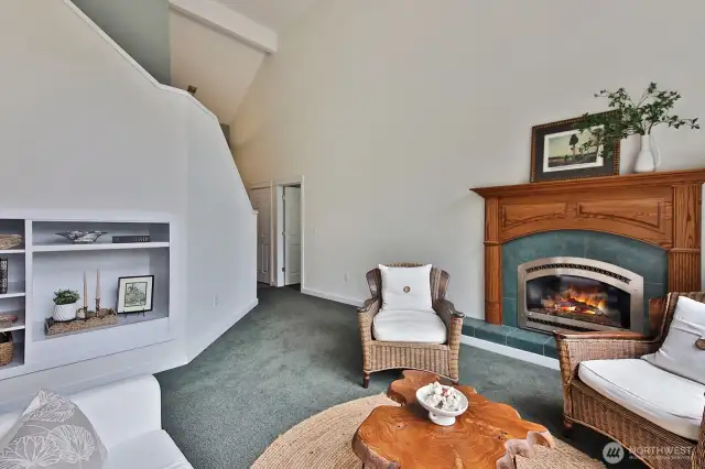 You are standing in the family room with cathedral ceiling, propane fireplace and built in shelving along the staircase. The open door in the distance is the Primary Suite.