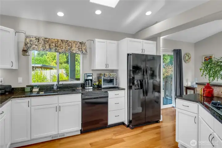 Beautiful functional kitchen featuring granite countertops, white cabinetry, and stainless appliances. A skylight and large window bring in abundant natural light, with easy access to the dining area and covered deck.
