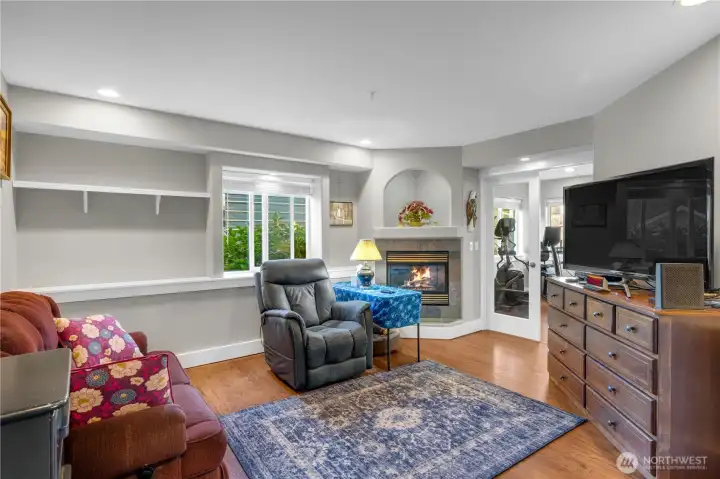 This family room features a gas fireplace, wood-style flooring, and soft natural light. A comfortable space for relaxing, movie nights, or quiet reading by the fire.