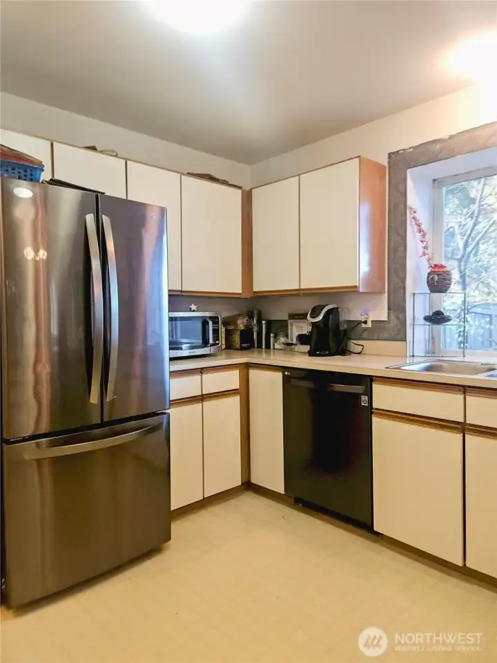 This angle showcases the roomy U-shaped layout, offering generous cabinet storage and ample counter space for cooking and coffee-making alike. The stainless-steel refrigerator and large window add both functionality and natural light, making the kitchen feel bright, practical, and inviting.