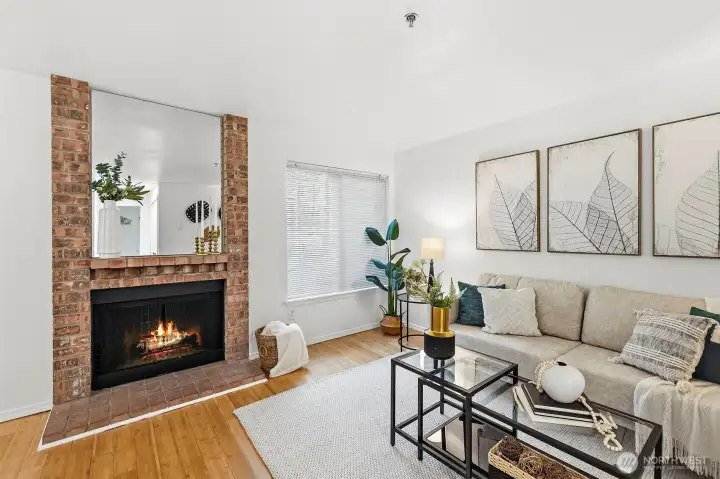 Inviting living room anchored by a classic brick wood-burning fireplace with mirror surround, hardwood floors, and large windows.