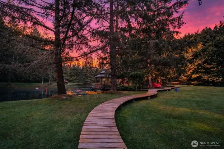 A raised wooden pathway meanders through the grounds, creating a quiet connection between the waterfront, guest quarters, wellness spa, and greenhouse. Framed by mature evergreens and open lawn, the boardwalk gives the property a resort-like sense of movement, discovery, and retreat.