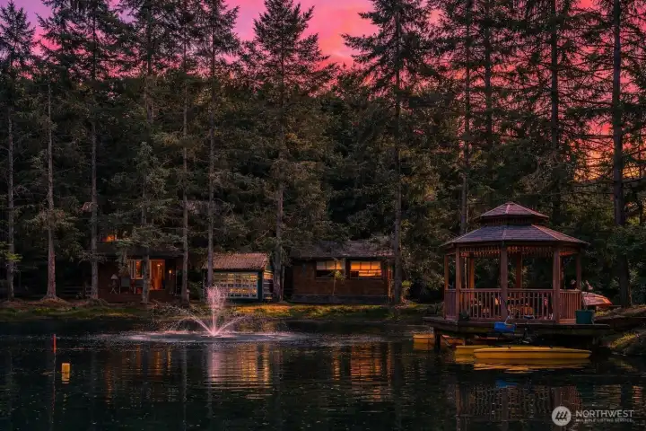 At dusk, the pond becomes a serene focal point with a fountain, waterside gazebo, and warm reflections from the surrounding guest accommodations. Framed by evergreens and soft evening light, the setting feels private, restorative, and quietly resort-like.
