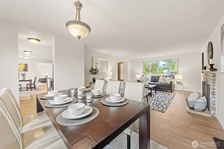 Open-concept dining and living area with large front-facing window and updated flooring throughout. Brick fireplace adds a central focal point to the inviting layout.