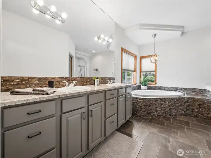 Refined spa-like bath featuring dual sinks, slab counters, a tiled corner soaking tub, and coordinated tile flooring. Refined spa-like bath featuring dual sinks, slab counters, a tiled corner soaking tub, and coordinated tile flooring.