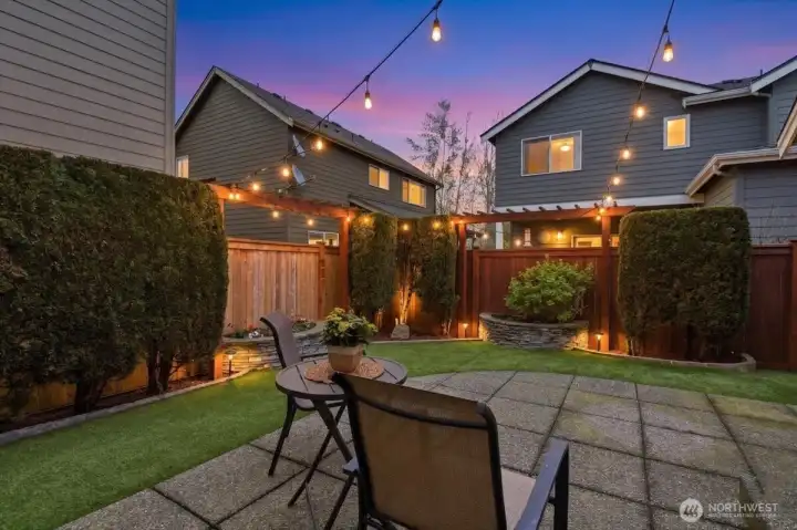 Twilight backyard patio scene with ambient string lights and fenced privacy, creating a warm outdoor entertaining atmosphere.