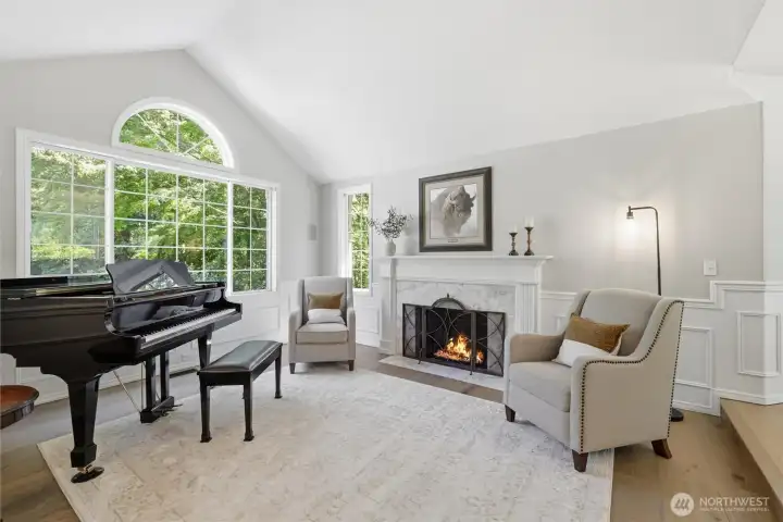 Stunning space to welcome guests or enjoy a quiet evening by the fire. This elegant formal living room showcases soaring vaulted ceilings, a dramatic arched picture window, rich millwork, and a beautiful wood-burning fireplace with a classic mantel surround. Bathed in natural light and framed by tranquil woodland views, the room offers a warm yet sophisticated atmosphere perfect for conversation, music, or relaxation. Thoughtful architectural details and timeless finishes create a truly memorable first impression.