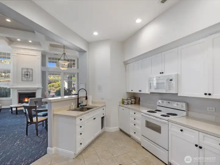 Community kitchen with cabinetry, sink, range, microwave, and open connection to clubhouse seating areas. Shared amenity space for gatherings and events.