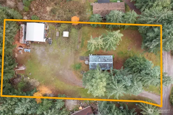 A drone view outlining the full 1.22-acre property, showing the home, the detached shop, and the wide open space between them. The lot is surrounded by tall evergreens for privacy and gives a great sense of the layout, usable areas, and separation between structures. This overhead view highlights how much room there is for parking, projects, and outdoor living.
