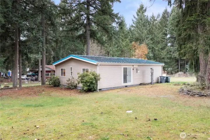 A view of the side and rear of the home, showing the level yard and the quiet forest backdrop. This angle highlights the sliding door leading to the main living area and gives buyers a sense of the home’s natural surroundings and the usable outdoor space.